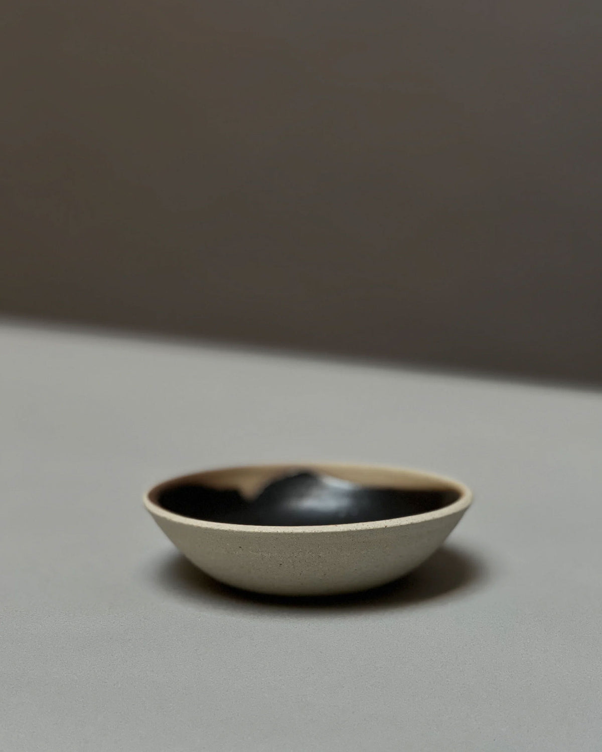 A round, bowl-like, ceramic incense holder with a natural unglazed outer surface and black glazed inner surface sits on a light grey table surface.