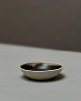 A round, bowl-like, ceramic incense holder with a natural unglazed outer surface and black glazed inner surface sits on a light grey table surface.
