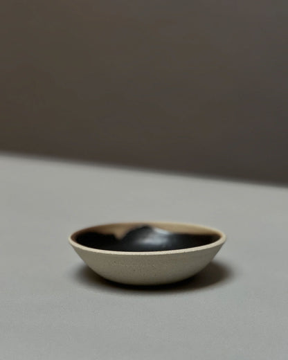 A round, bowl-like, ceramic incense holder with a natural unglazed outer surface and black glazed inner surface sits on a light grey table surface.