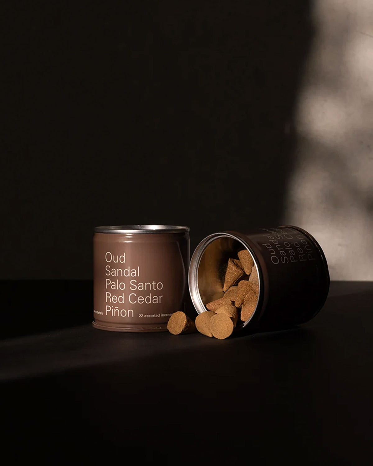A short brown tin can with white lettering lists Oud, Sandal, Palo Santo, Red Cedar, Piñon. A second tin is on its side with the lid removed, showing the contents of small brown incense cones spilling out onto a dark table surface against moody lighting and shadows. 