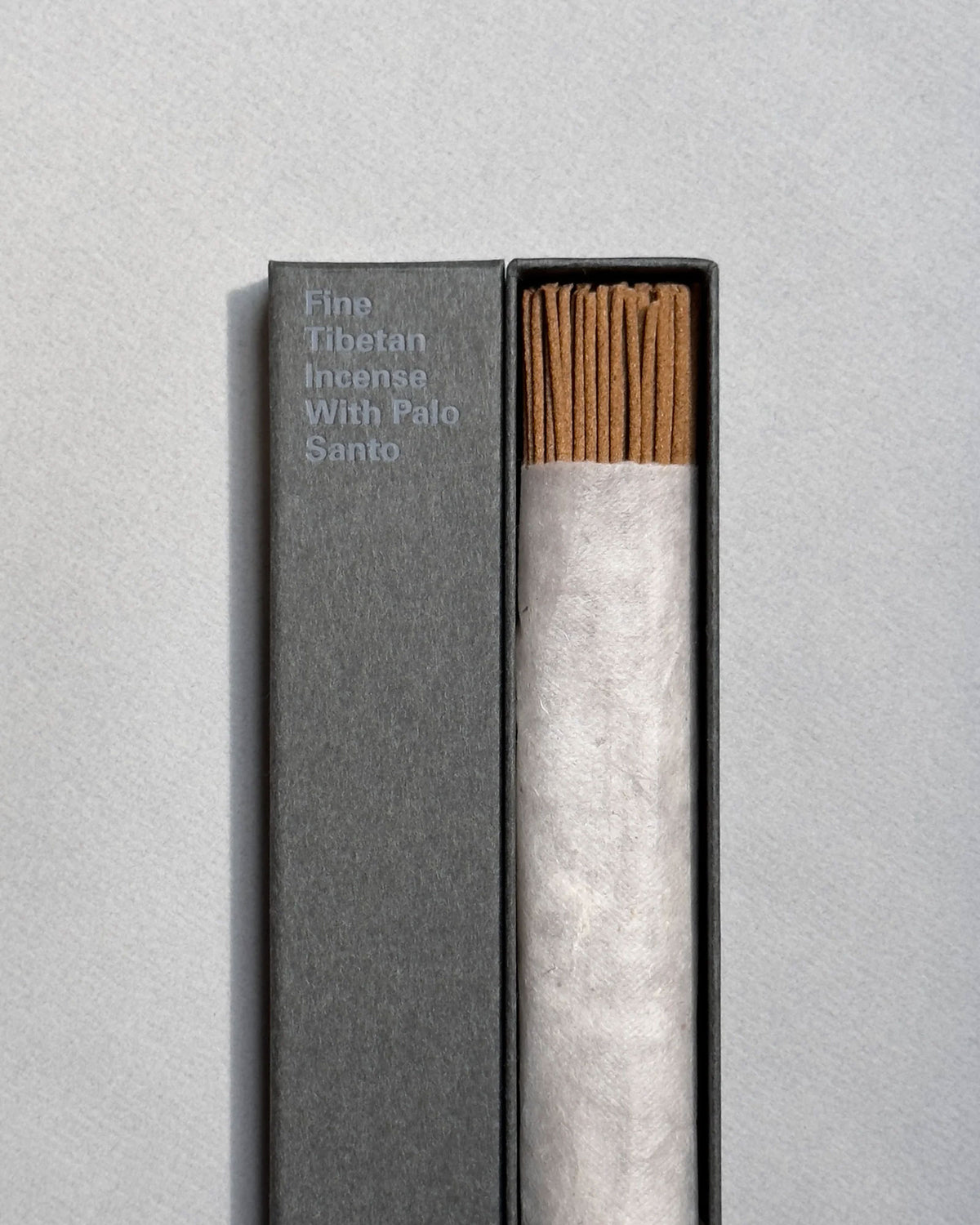 A long, rectangular, grey box is open to show a wrapped bundle of light brown incense sticks, the lid of the same colour sits next to the incense. On the front of the lid in light blue printed lettering, it reads Fine Tibetan Incense With Palo Santo. Set on a seamless light grey background. 