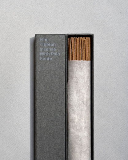 A long, rectangular, grey box is open to show a wrapped bundle of light brown incense sticks, the lid of the same colour sits next to the incense. On the front of the lid in light blue printed lettering, it reads Fine Tibetan Incense With Palo Santo. Set on a seamless light grey background. 