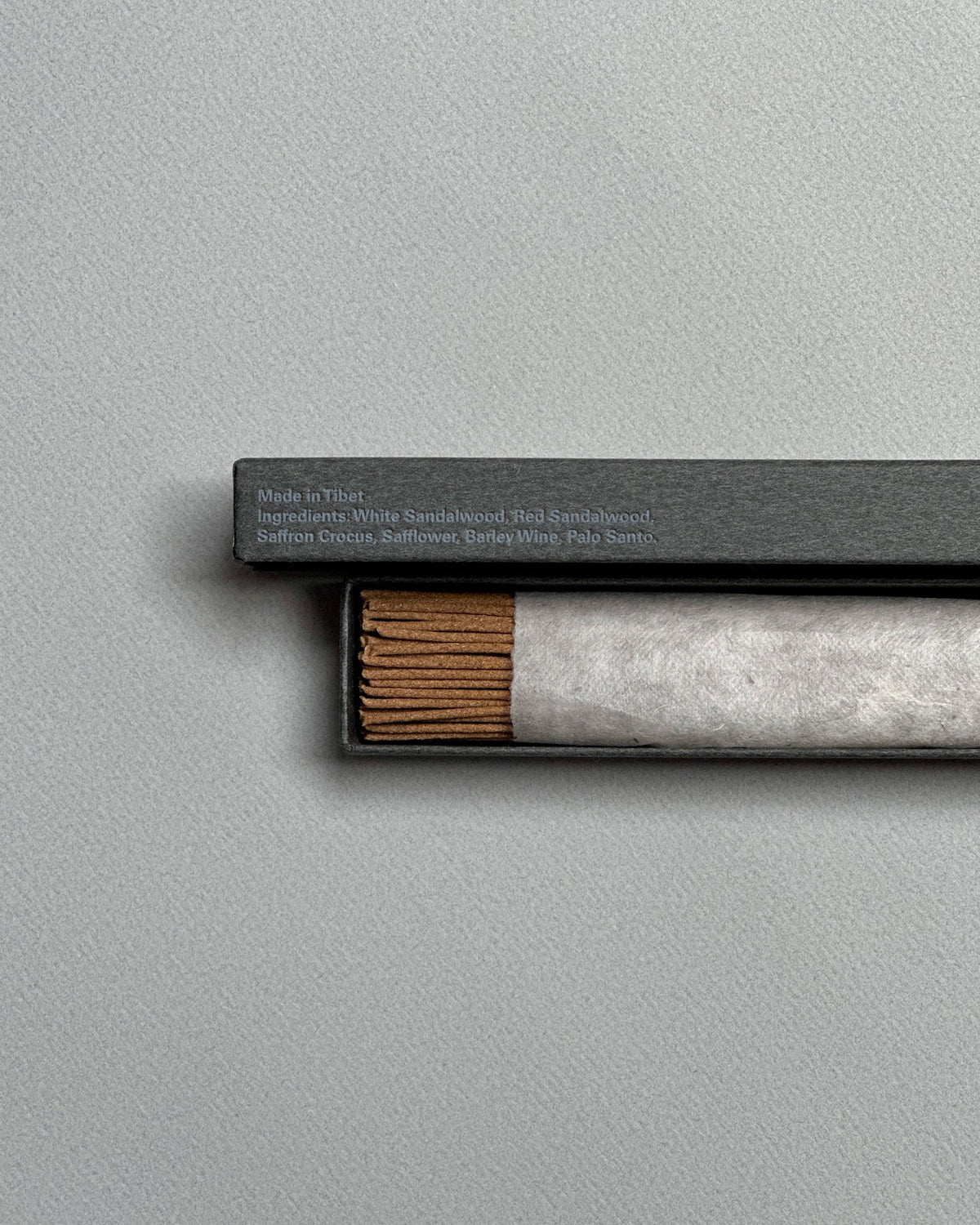 A long, rectangular, grey box is open to show a wrapped bundle of light brown incense sticks, the lid of the same colour sits above the incense. On the side of the lid in light blue printed lettering, it reads Made in Tibet, White sandalwood, Red Sandalwood, Saffron Crocus, Safflower, Berley Wine, Palo Santo. Set on a seamless light grey background. 