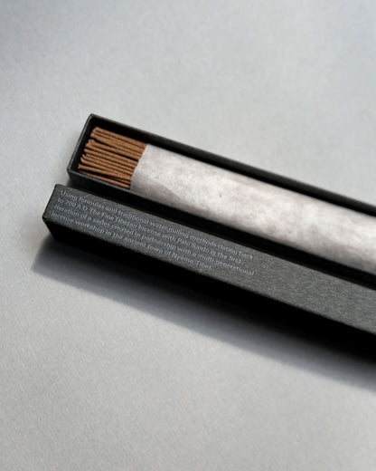 A long, rectangular, grey box is open to show a wrapped bundle of light brown incense sticks, the lid of the same colour sits below the incense. On the side of the lid in light blue printed lettering is a paragraph of text. Set on a seamless light grey background. 