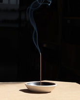 A tall, light brown incense stick is sitting straight up from the centre of a round ceramic dish with a matte white outer finish and satin black inner finish. The incense is lit, and a line of smoke drifts from the tip of the incense. Set on a light table surface with a dark, almost black background. 