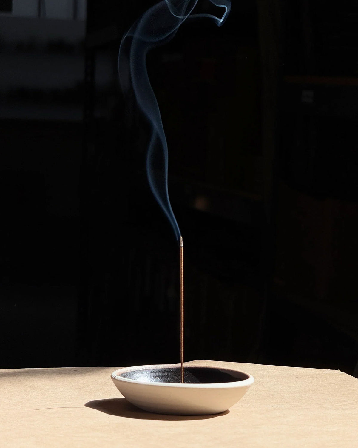 A tall, light brown incense stick is sitting straight up from the centre of a round ceramic dish with a matte white outer finish and satin black inner finish. The incense is lit, and a line of smoke drifts from the tip of the incense. Set on a light table surface with a dark, almost black background. 