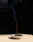 A tall, light brown incense stick is sitting straight up from the centre of a round ceramic dish with a matte white outer finish and satin black inner finish. The incense is lit, and a line of smoke drifts from the tip of the incense. Set on a light table surface with a dark, almost black background. 