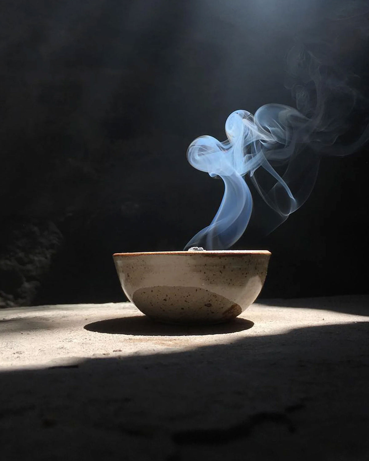 A speckled ceramic bowl sits on a dark surface with dramatic sunlight and a beam of light shining down, creating a plume of white and blue smoke rising from inside the bowl.