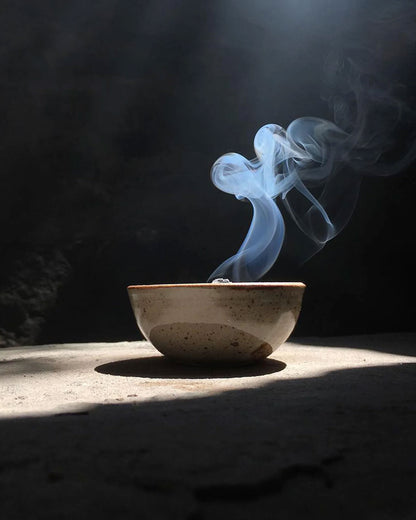 A speckled ceramic bowl sits on a dark surface with dramatic sunlight and a beam of light shining down, creating a plume of white and blue smoke rising from inside the bowl.