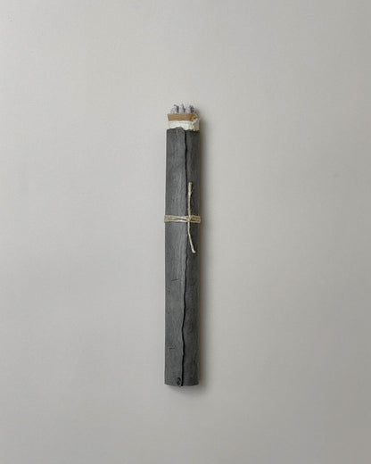 A bundle of grey coloured Copal incense sticks wrapped in layers of paper, the final layer is a dark grey, almost blue, silky textured paper, plain string is tied around the centre, only the very tips of the incense are visible. Set on a light grey background. 