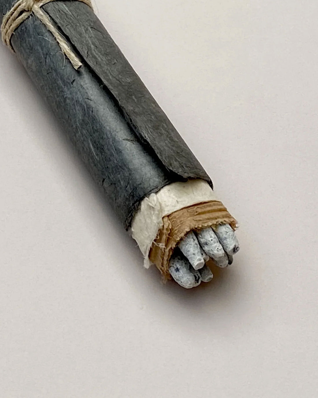 A bundle of grey coloured Copal incense sticks wrapped in layers of paper, the final layer is a dark grey, almost blue, silky textured paper, plain string is tied around the centre, only the very tips of the incense are visible. Set on a light grey background. 