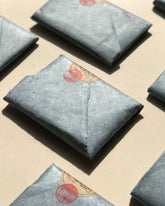 Several rectangular blue-grey wrapped parcels arranged on a beige surface, with one centered in the foreground and partial duplicates visible around the edges. The center item has a small red circular stamp and round brown sticker with the Incausa logo.