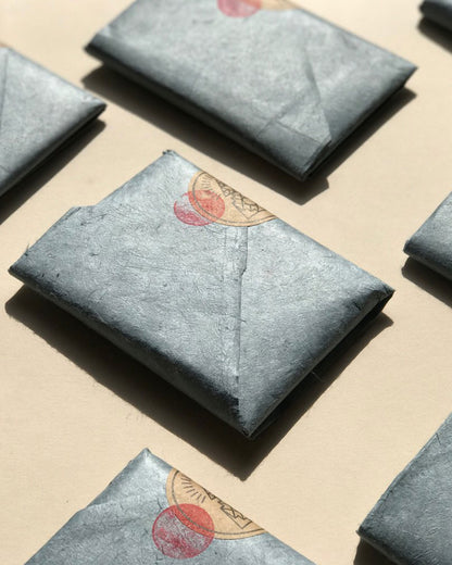 Several rectangular blue-grey wrapped parcels arranged on a beige surface, with one centered in the foreground and partial duplicates visible around the edges. The center item has a small red circular stamp and round brown sticker with the Incausa logo.