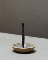 A single dark charcoal incense stick stands upright in a shallow, textured ceramic incense holder with a warm beige exterior and a gray interior, set against a minimalist two-tone gray background.