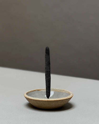 A single dark charcoal incense stick stands upright in a shallow, textured ceramic incense holder with a warm beige exterior and a gray interior, set against a minimalist two-tone gray background.