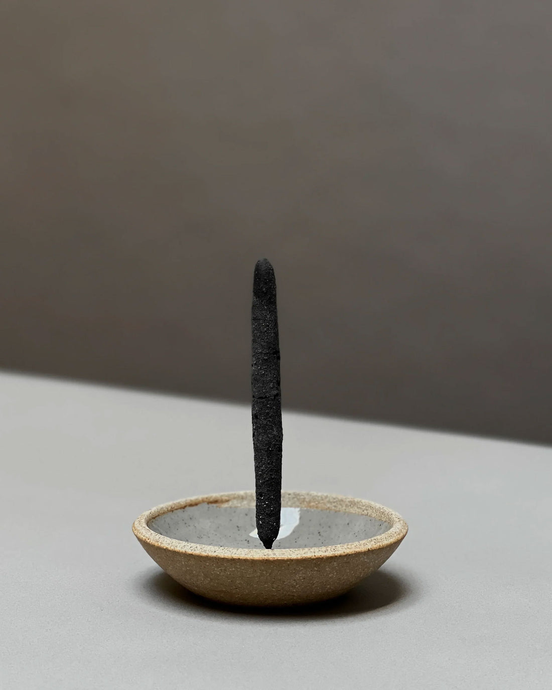 A single dark charcoal incense stick stands upright in a shallow, textured ceramic incense holder with a warm beige exterior and a grey interior, set against a minimalist two-tone grey background.