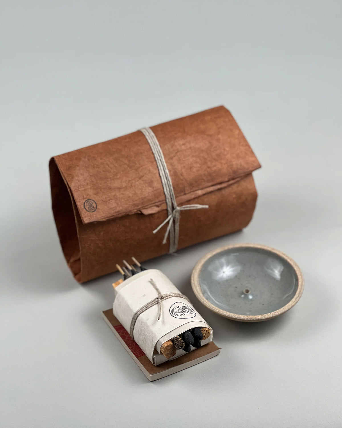 A product shot showing the Incausa Home Bundle brown paper wrapped parcels along with its contents of incense and incense holder. Set on a seamless, light grey background.