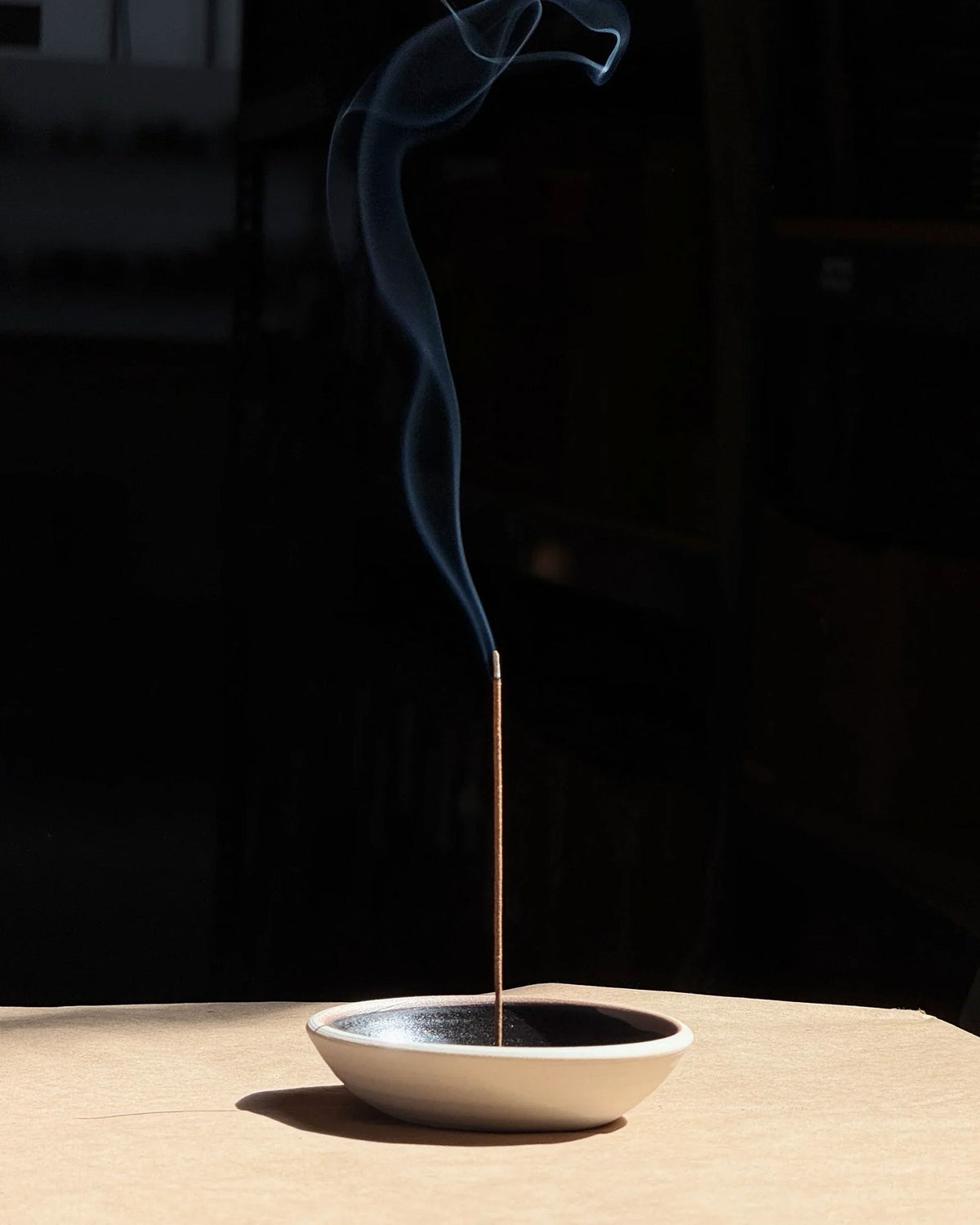 A single smouldering brown incense stick stands upright in a shallow, textured ceramic incense holder with a warm beige exterior and a black interior, set against a minimalist two-tone grey background.