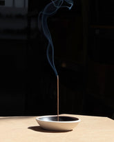 A single smouldering brown incense stick stands upright in a shallow, textured ceramic incense holder with a warm beige exterior and a black interior, set against a minimalist two-tone grey background.