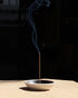 A single smouldering brown incense stick stands upright in a shallow, textured ceramic incense holder with a warm beige exterior and a black interior, set against a minimalist two-tone grey background.