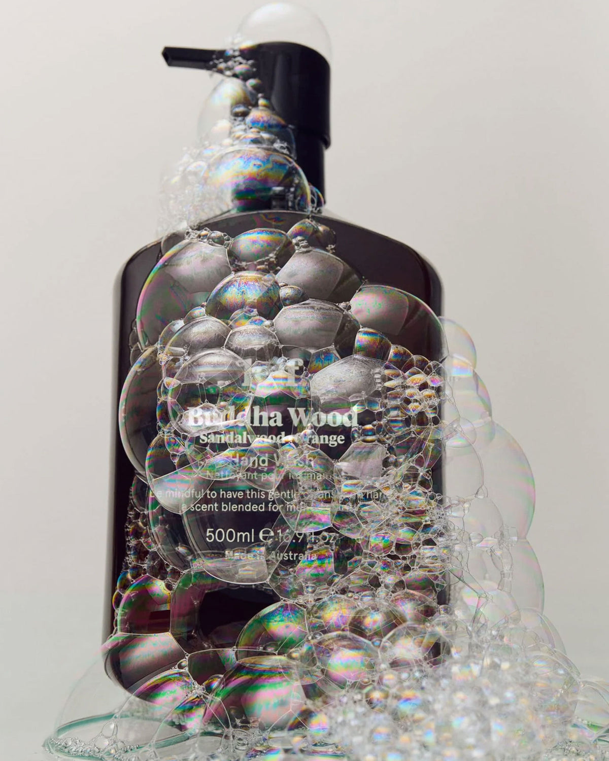 A 500ml dark amber bottle of Leif Buddha Wood Hand Cleanser with a black pump dispenser covered with soap bubbles against a plain grey background. 