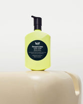 Leif Desert Lime Body Lotion in a bright yellow bottle with a black pump and black circular label, displayed on a cream-coloured sculptural surface against a white background.
