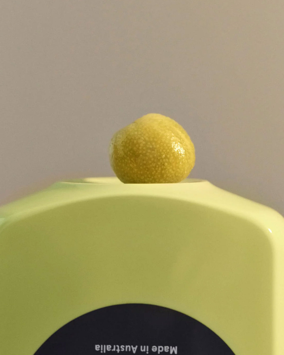 A close-up of a bright yellow bottle with a small yellow citrus fruit resting on top, against a plain light grey background. A black circular label at the bottom reads &