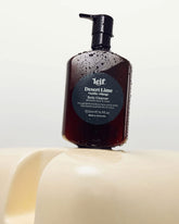 A 500ml dark amber bottle of Leif Desert Lime Body Cleanser with a black pump dispenser on a cream coloured sculptural surface.