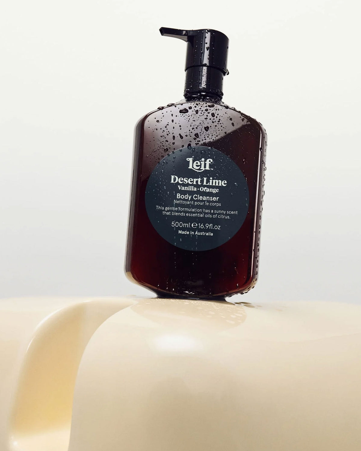 A 500ml dark amber bottle of Leif Desert Lime Body Cleanser with a black pump dispenser on a cream coloured sculptural surface.