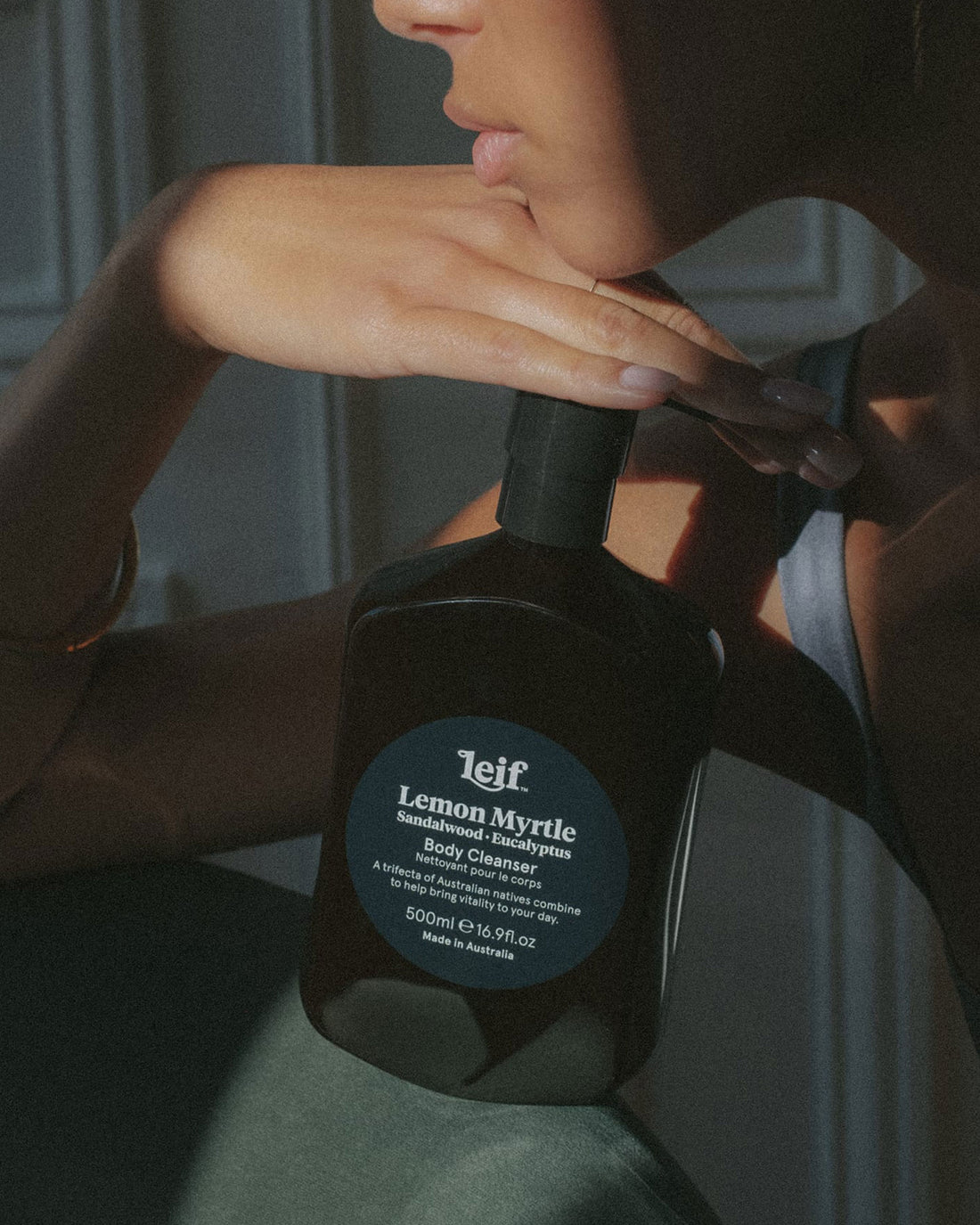 A 500ml dark amber bottle of Leif Lemon Myrtle Body Cleanser with a black pump dispenser, resting on a chair with a woman&