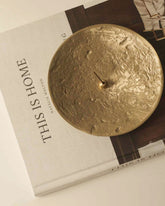 A round brass textured plate sits on top of a large coffee-table book titled This is Home on an off-white table surface. In the centre of the brass plate is a small brass incense holder with an incense stick.