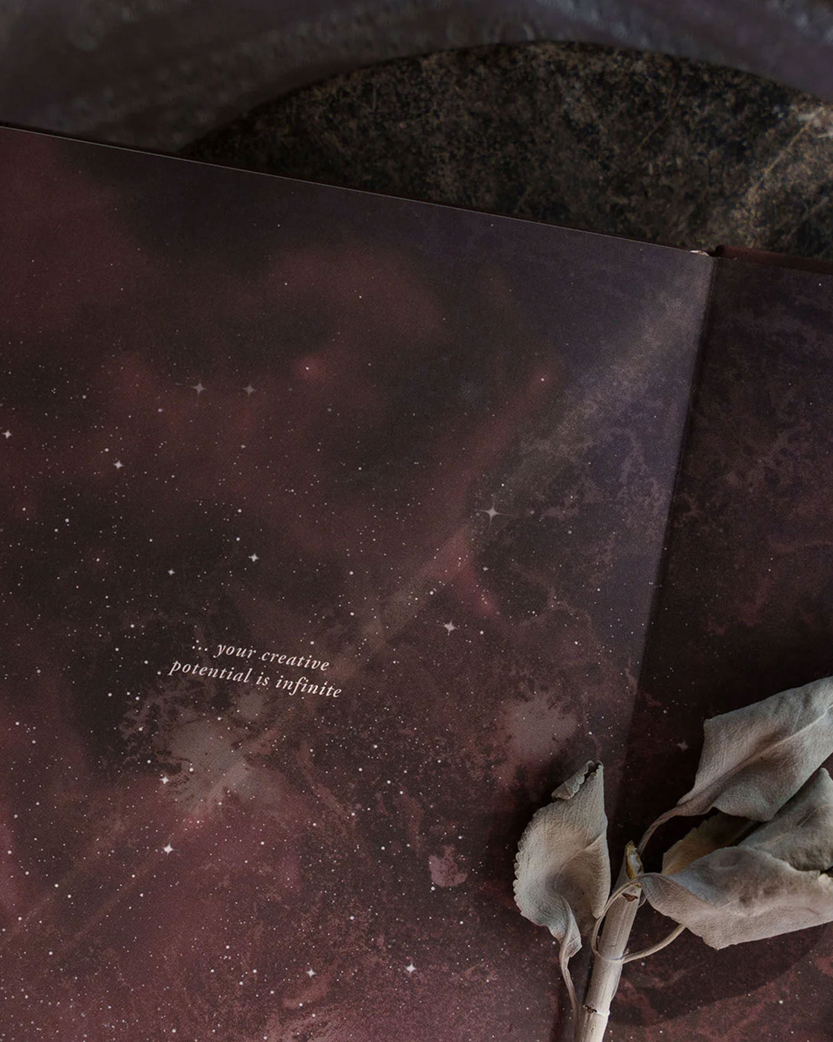 An open book with a dark purple starry night background and centred text that reads "...your creative potential is infinite". A sprig of dried white sage is placed in the fold of the open book. 