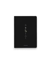 A black hardcover notebook featuring the Capricorn zodiac sign and its chart symbols printed in metallic on the front cover, set against a clean white background.