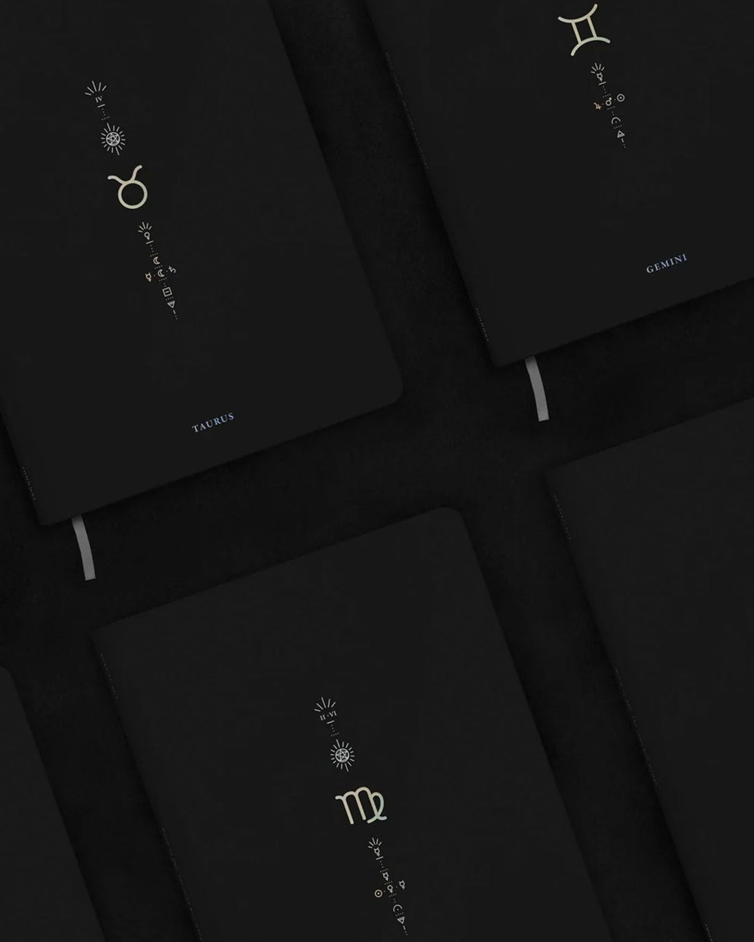 A close-up, angled view of several black hardcover notebooks arranged on a dark surface. Each notebook features a different zodiac sign printed in metallic gold, including Taurus, Gemini and Virgo.