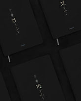 A close-up, angled view of several black hardcover notebooks arranged on a dark surface. Each notebook features a different zodiac sign printed in metallic gold, including Taurus, Gemini and Virgo.