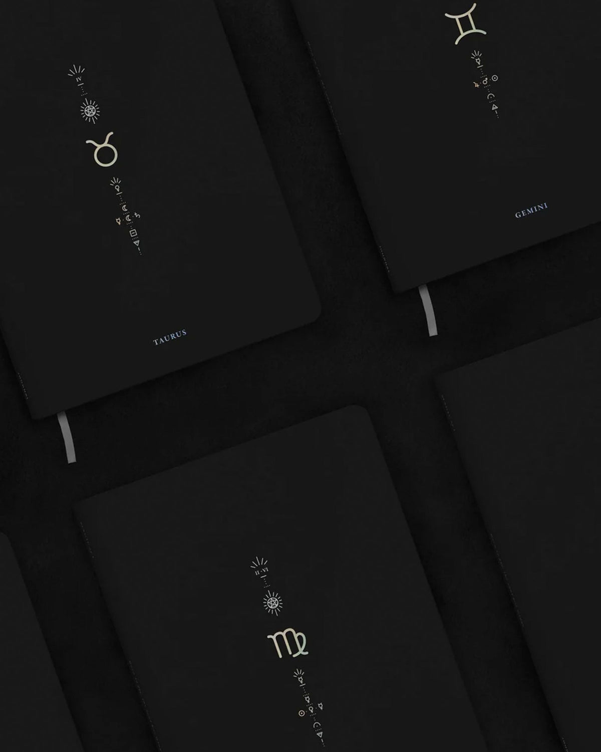 A close-up, angled view of several black hardcover notebooks arranged on a dark surface. Each notebook features a different zodiac sign printed in metallic gold, including Taurus, Gemini and Virgo.
