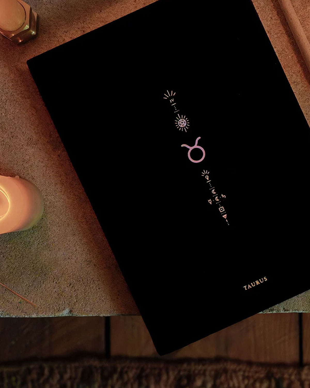 A black journal with Taurus zodiac symbols photographed at an angle on a textured surface with a warm candle nearby.