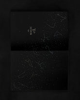 A black hardcover notebook or journal with a starry constellation pattern, shown open against a dark background. The top cover features the text &