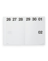 A white paper calendar page spread open, showing the days of the week with black printed numbers. The page is photographed against a clean white background.