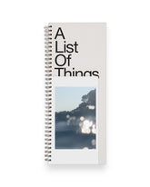 A tall ledger style notebook with wire binding, cover reads in large black letters A List Of Things with a postcard style picture of sun glare on water. Set on a solid white background.