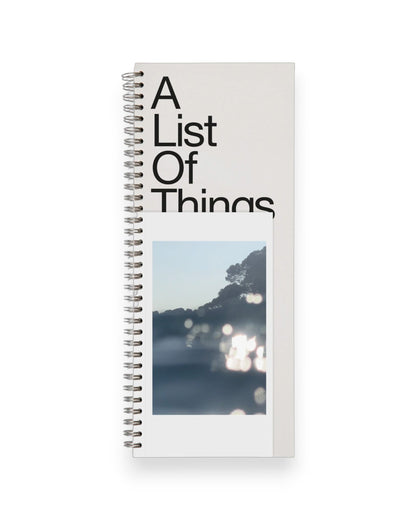 A tall ledger style notebook with wire binding, cover reads in large black letters A List Of Things with a postcard style picture of sun glare on water. Set on a solid white background.