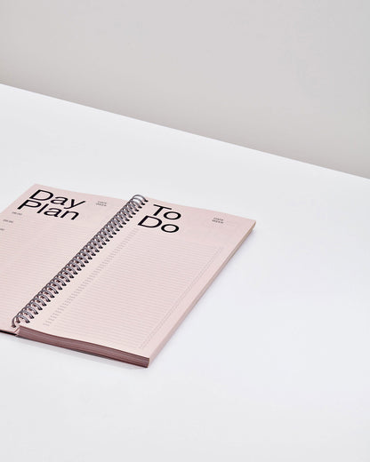 An open tall, rectangular ring-bound planner with light pink pages on a white table surface.
