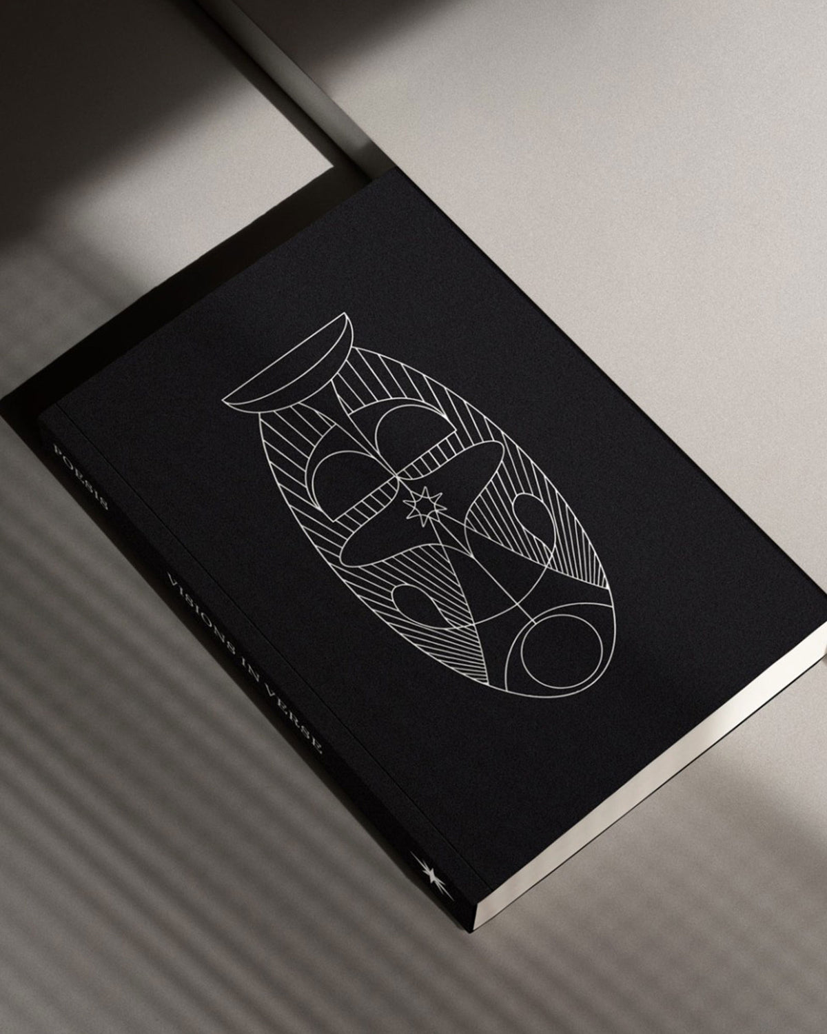 A copy of The Poesis Visions and Verses Guidebook, an all-black cover with a white geometric illustration. Set on a white table surface with dramatic shadows.