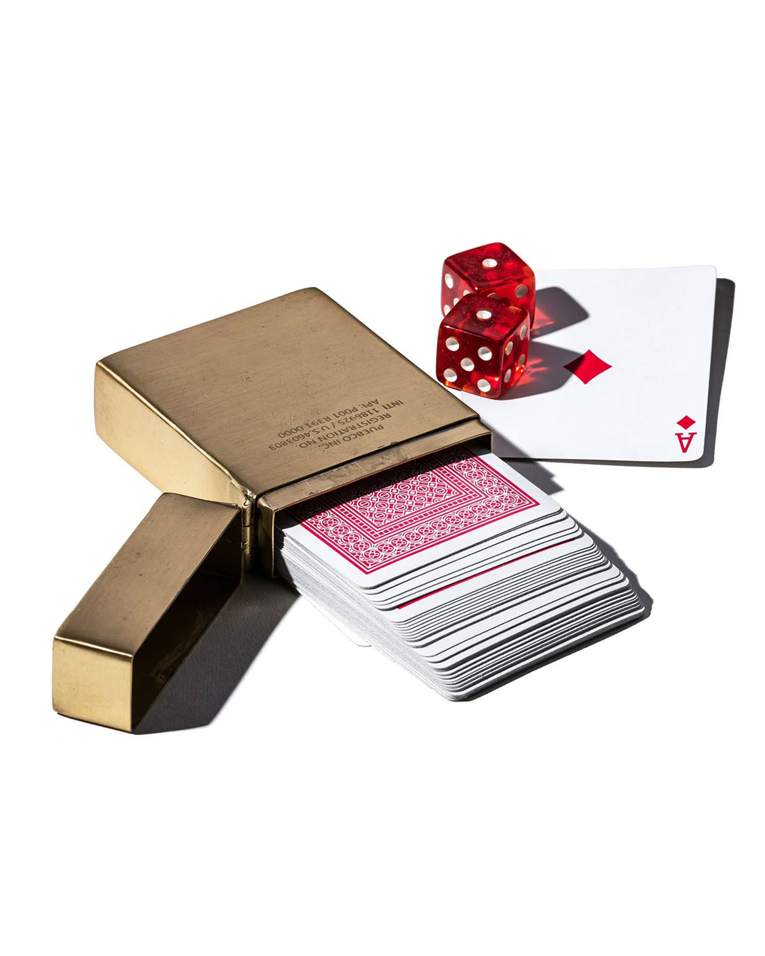 An open rectangular brass card case with a brushed satin finish shows a full deck of playing cards. An ace of diamonds and two red dice are next to the card case. Set on white background.