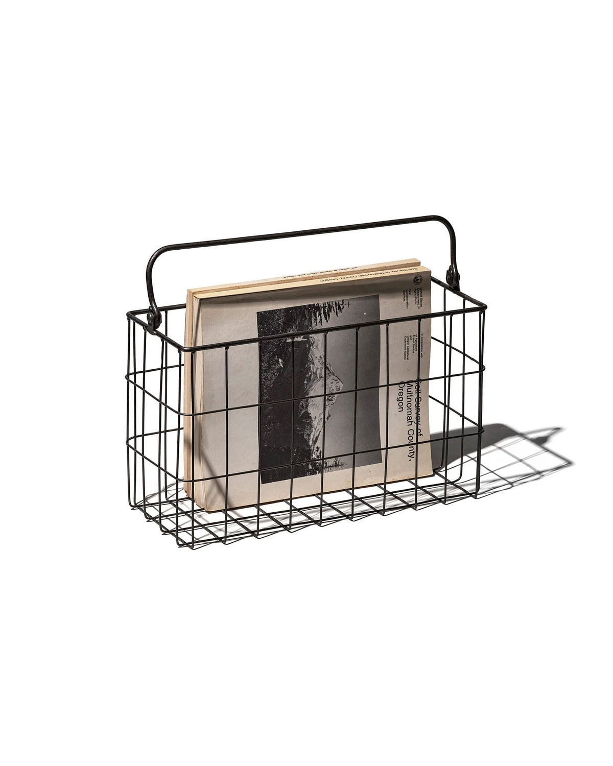 A black rectangular wire basket with handle holding several black and white magazines. Set on a white background. 