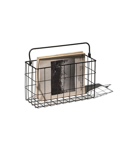 A black rectangular wire basket with handle holding several black and white magazines. Set on a white background. 