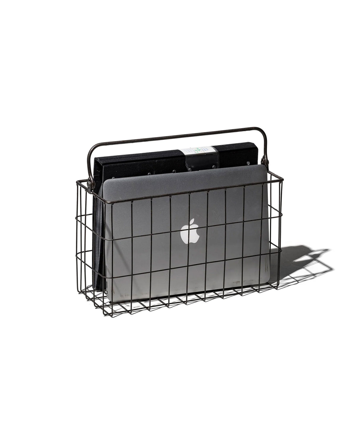 A black rectangular wire basket with handle holding a mac lap book and a black folder. Set on a white background. 