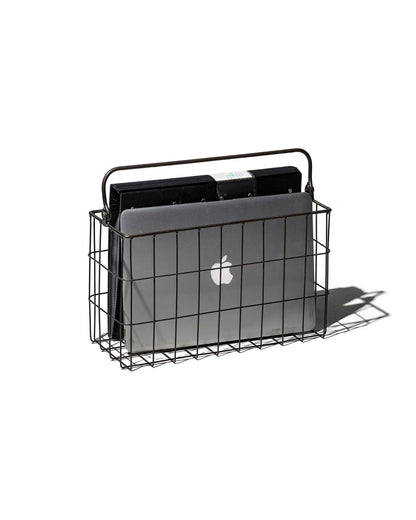 A black rectangular wire basket with handle holding a mac lap book and a black folder. Set on a white background. 