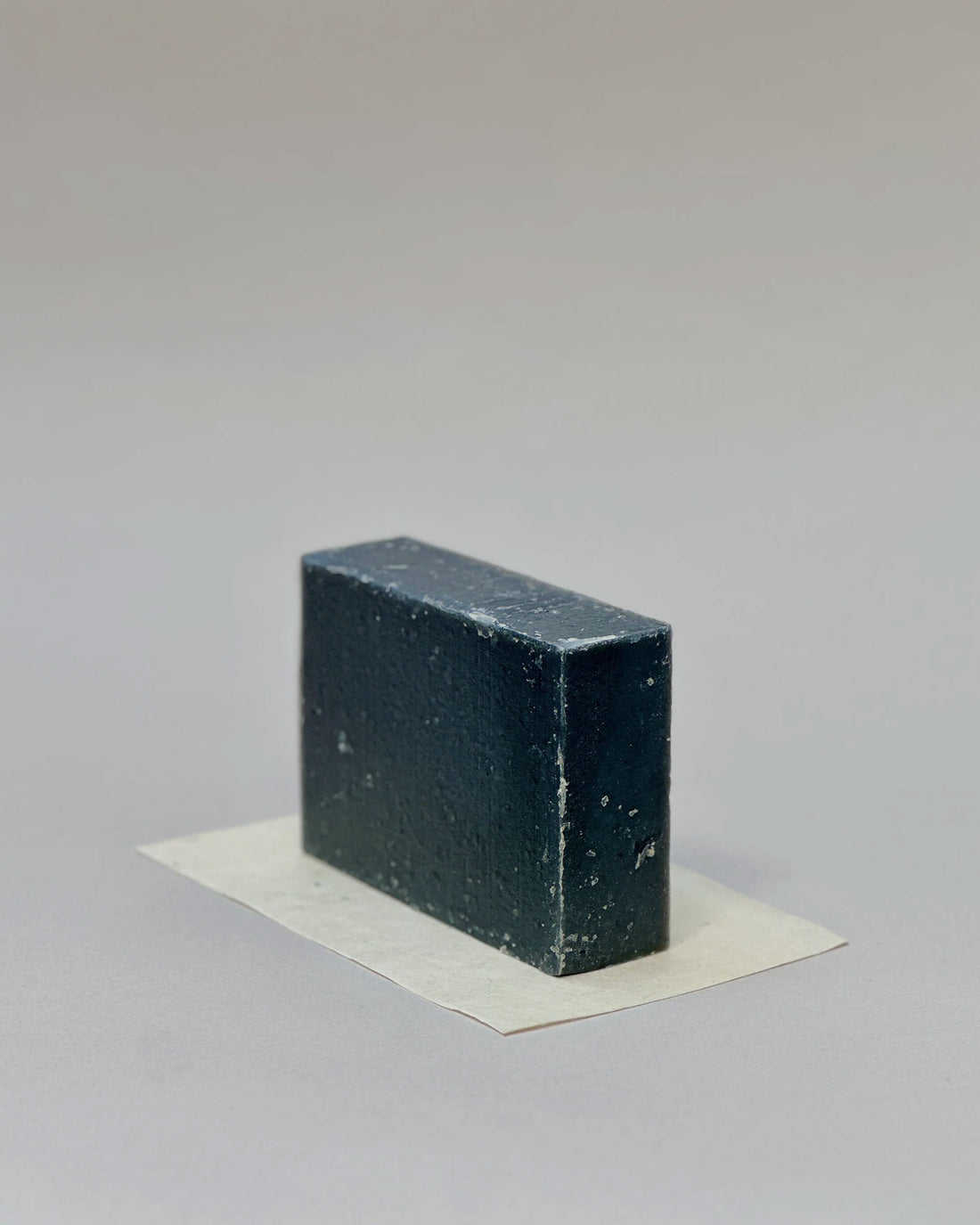 A rectangular bar of charcoal soap sitting on its side on a rectangular piece of white paper against a seamless, light grey background. 
