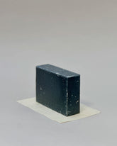 A rectangular bar of charcoal soap sitting on its side on a rectangular piece of white paper against a seamless, light grey background. 